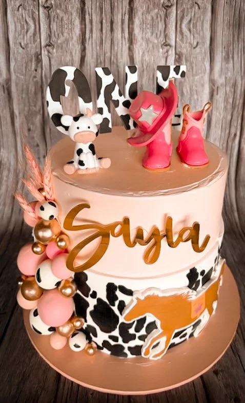 A two-tiered cake with cow print and brown, pink, and white decorations, featuring cow and horse toppers, with the name 'Gaya' in gold on the front, set against a wooden background.