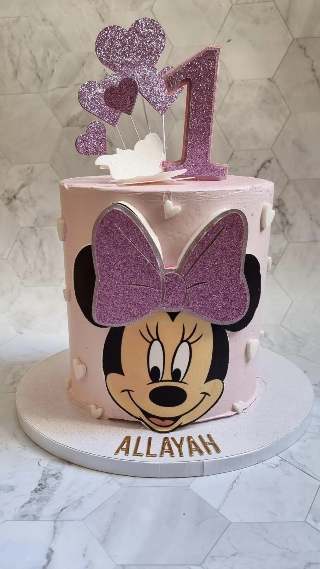 Pink birthday cake with a Minnie Mouse face, a large pink glitter bow, and a big pink glitter number one on top. The cake has white heart decorations and white gloves, with the name 'ALLAYAH' in gold letters at the bottom.