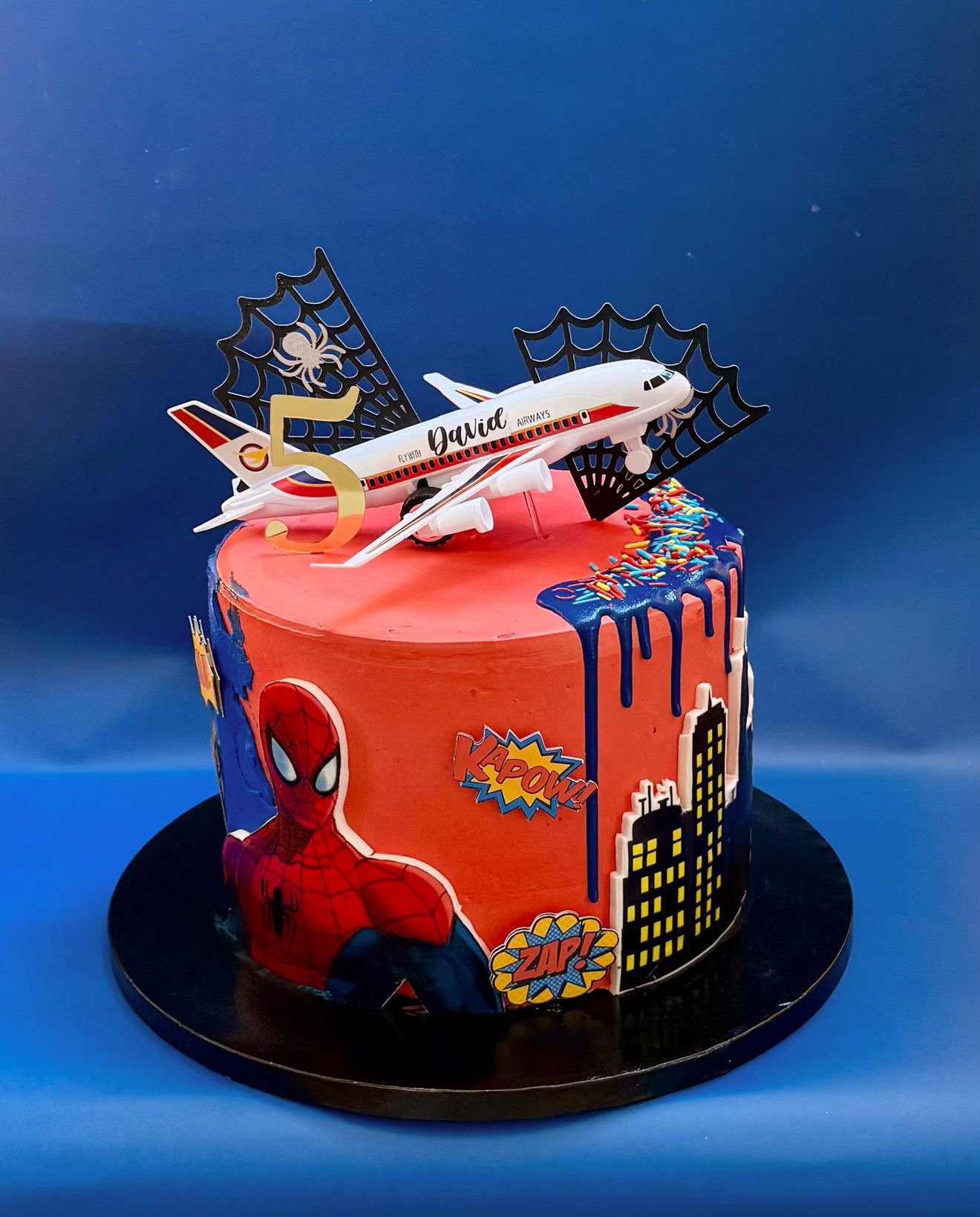 A Spider-Man themed birthday cake with superhero and comic book decorations, a toy airplane with Spider-Man branding on top, and a large gold number 5.