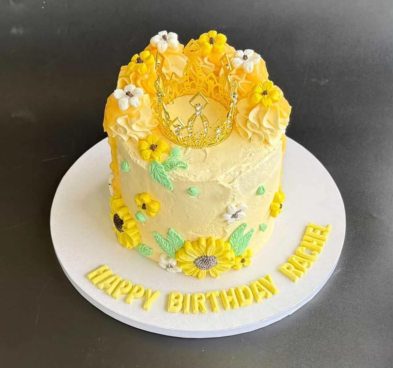 A birthday cake decorated with yellow and white flowers, a golden crown on top, and yellow letters spelling "HAPPY BIRTHDAY PRINCE" around the base.