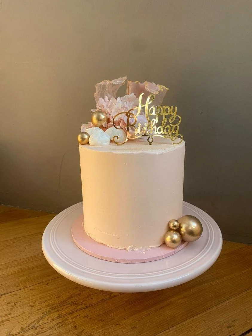 A round birthday cake with pale pink frosting, decorated with gold and white balls, pink flowers, and a gold "Happy Birthday" topper, placed on a white plate on a wooden surface.