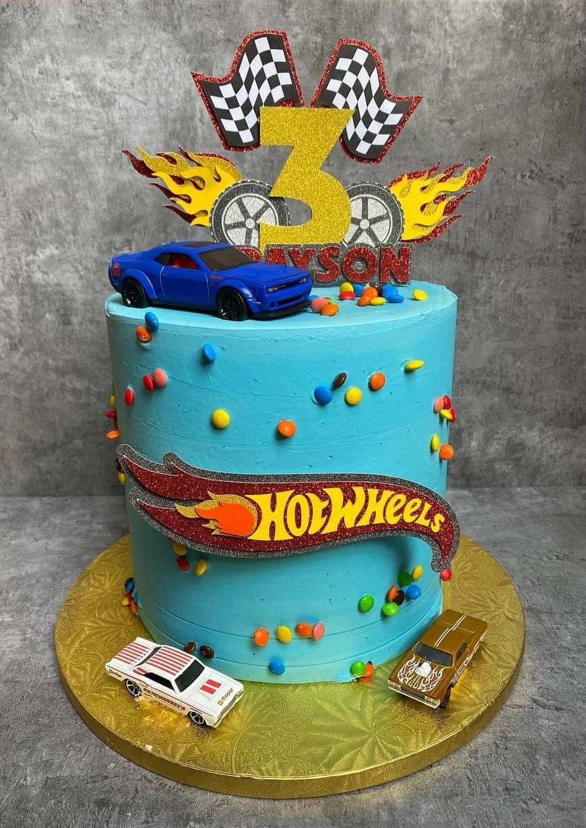 A blue Hot Wheels-themed birthday cake with colorful sprinkles, toy cars, checkered flags, and a large glittery yellow number 3 on top, celebrating a child's third birthday.
