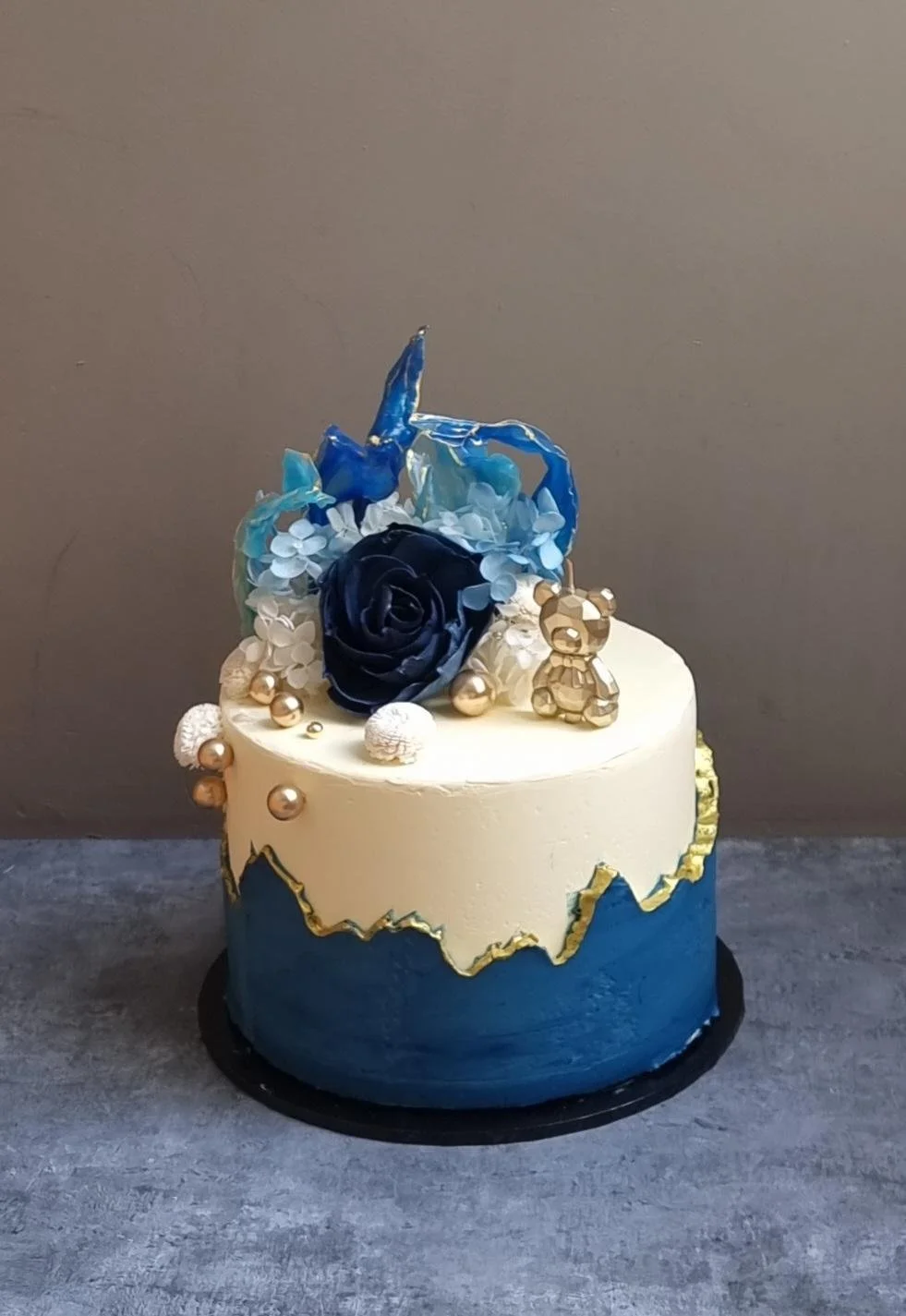 A decorated cake with white and blue icing, gold and pearl accents, flowers, a gold teddy bear, and blue and white decorative sugar elements on top.