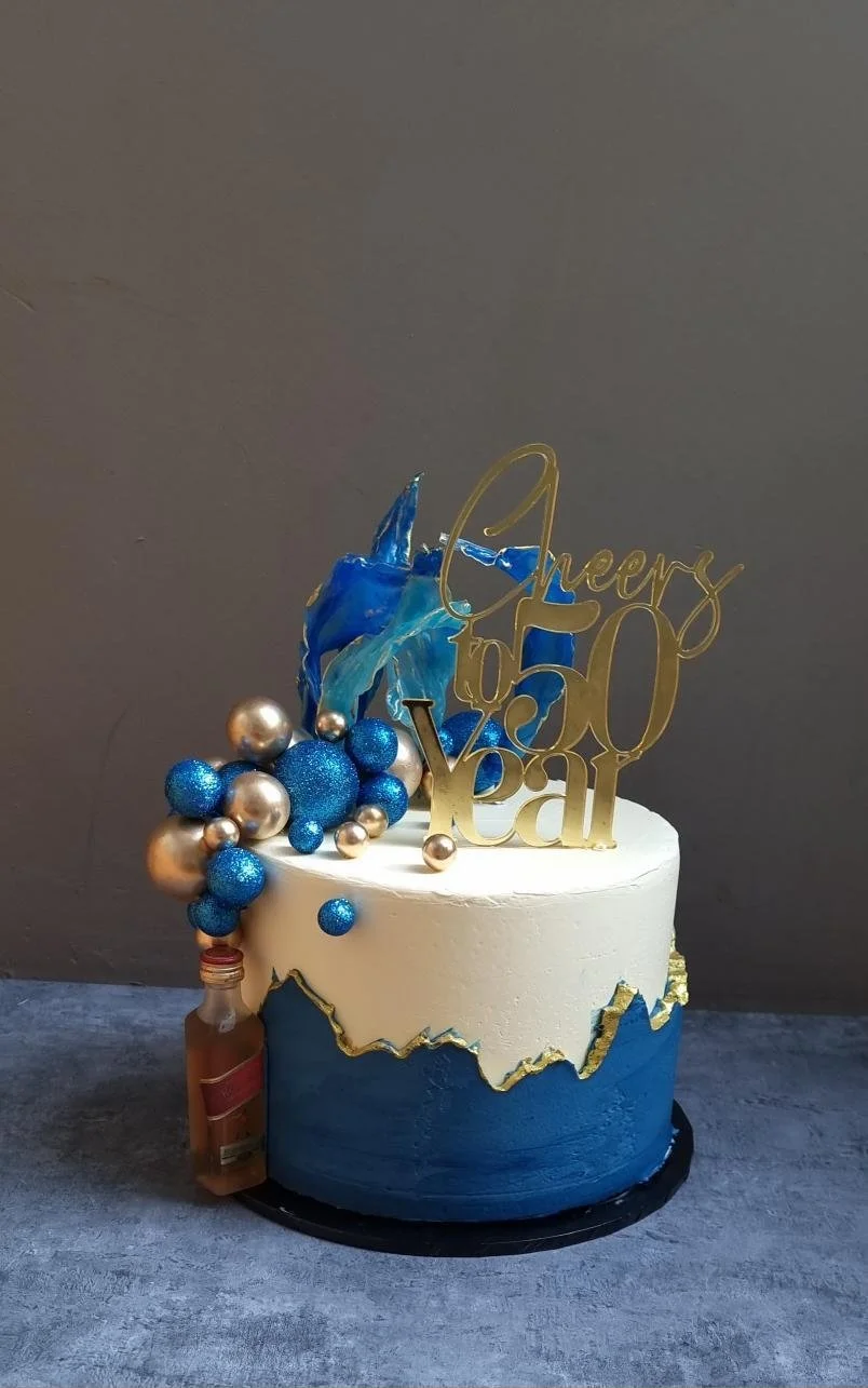 Celebratory cake with blue and white frosting, gold and blue decorations including a bottle, faux blue flames, and a gold topper that reads 'Cheers to 50 Year' on a gray background.