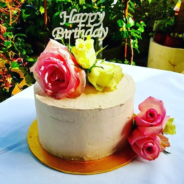 A round birthday cake with light pink frosting, decorated with pink and yellow roses, a 'Happy Birthday' topper, and some roses on a white tablecloth outside with green foliage in the background.