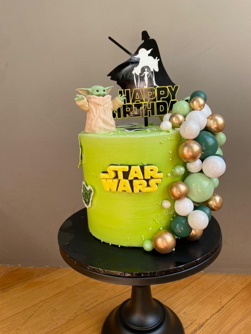 Star Wars themed birthday cake with green frosting, featuring a Baby Yoda figure, Star Wars logo, and a silhouette topper of Darth Vader with a lightsaber, decorated with gold, white, and green balloons.
