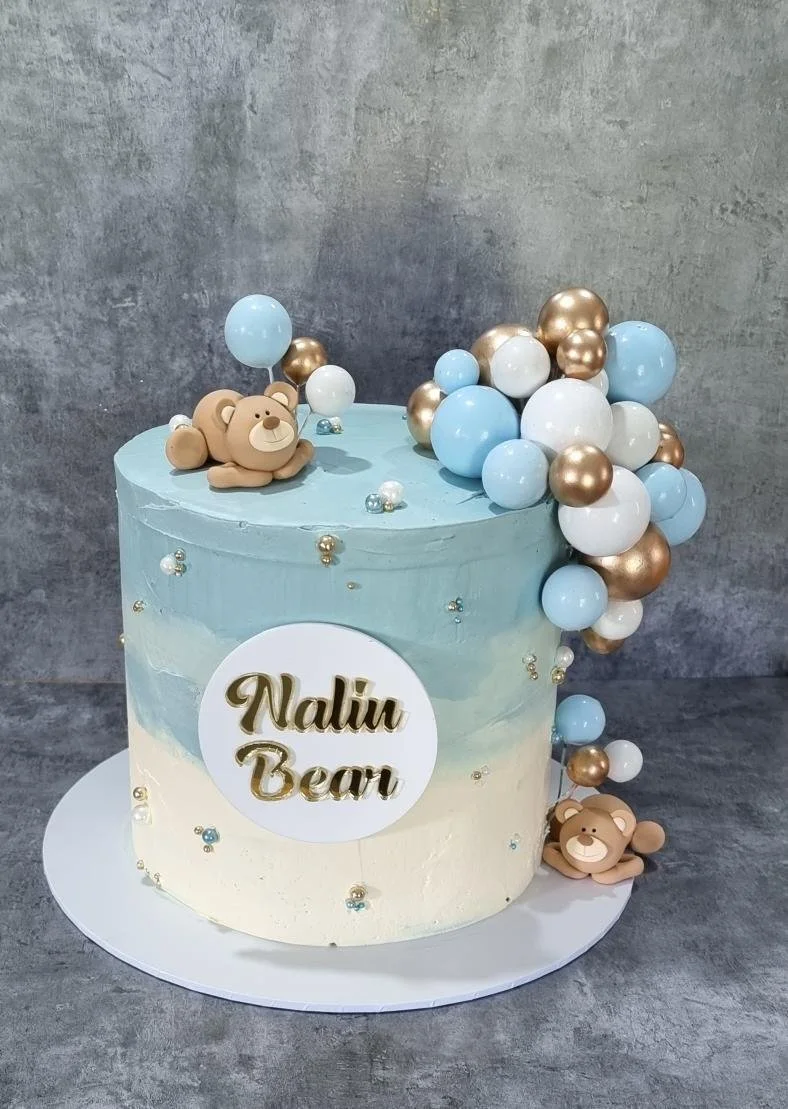 A two-layer cake with a blue and white gradient frosting, decorated with small pearl-like beads, featuring a bear figurine on top and a cluster of blue, white, and gold balloons cascading down one side. The cake has a white circle with the text 'Nati