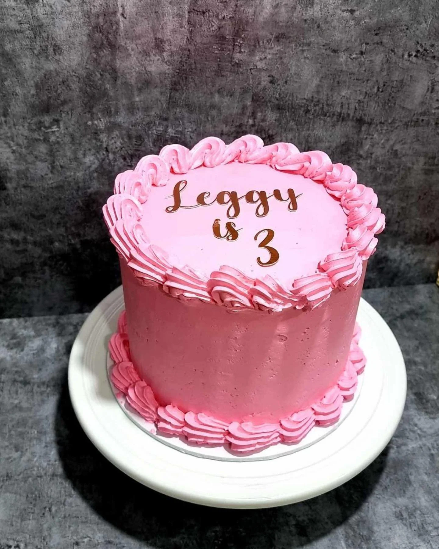 Pink birthday cake with decorative icing and a message reading "Leggy is 3" on top, placed on a white cake stand on a dark countertop.