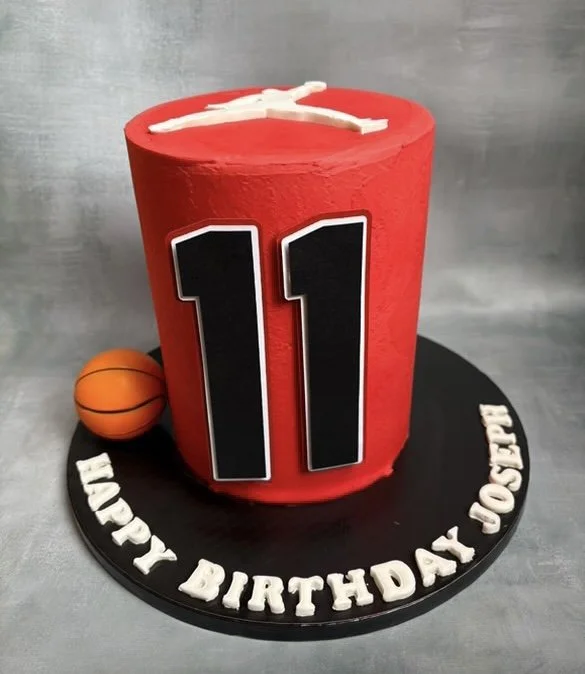 A red birthday cake with a white number 11 on the front, decorated with a basketball on the side, sitting on a black round cake base with white fondant letters spelling 'HAPPY BIRTHDAY JAXX JOSPH,' against a gray background.