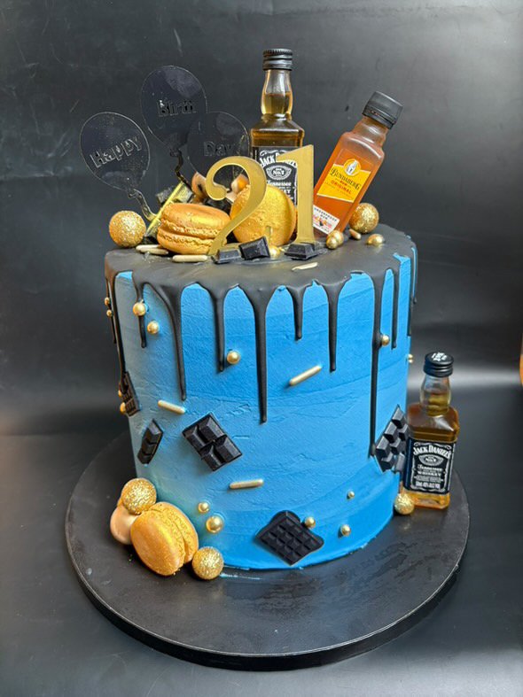 A blue birthday cake decorated with gold and black accents, including miniature bottles of Jack Daniel's whiskey, macarons, chocolate pieces, and gold spheres. The top of the cake features decorative elements such as macarons, gold balls, and whiskey