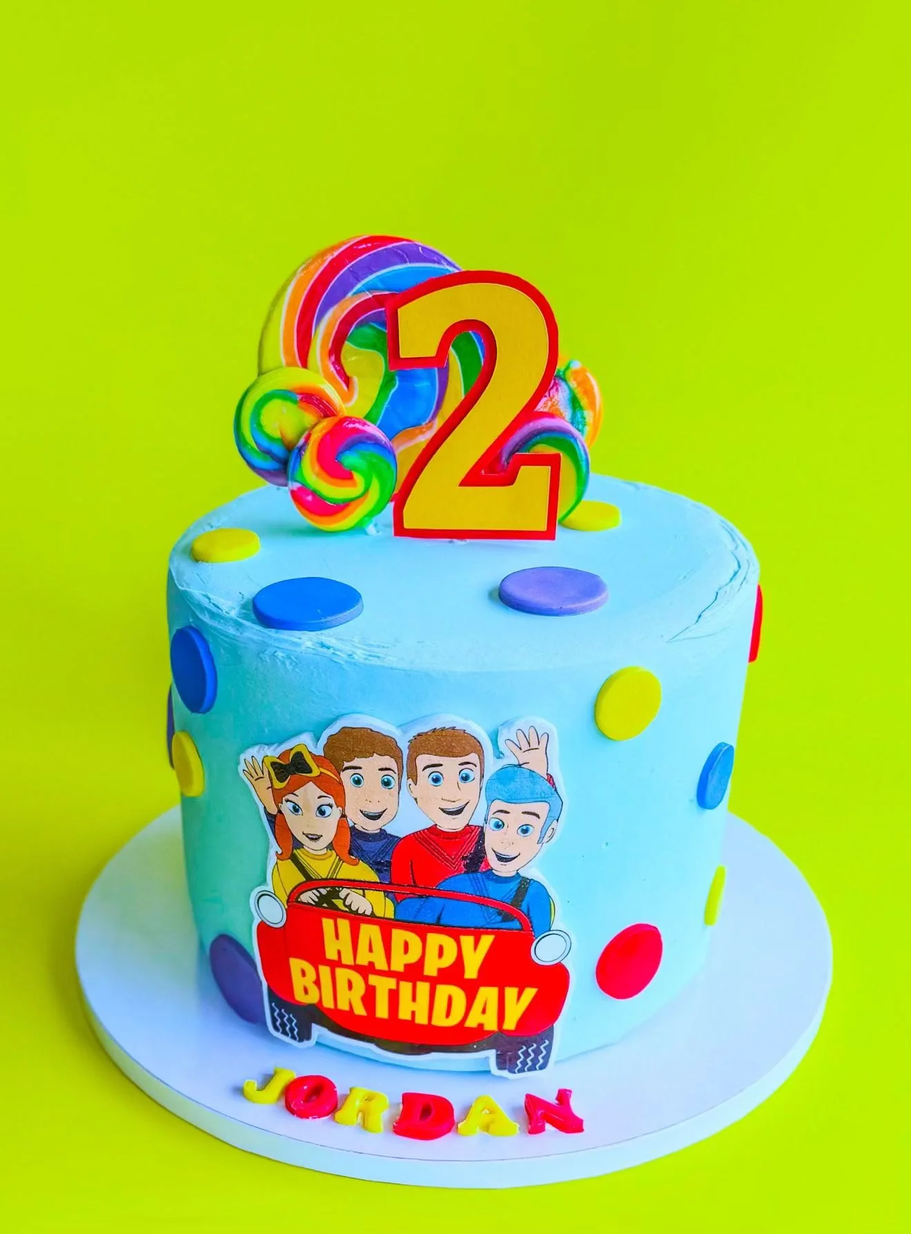 Colorful birthday cake with rainbow lollipops, circular candies, and a large red and yellow number 2 candle on top. The cake features a 'Happy Birthday' sticker with cartoon characters. Bright green background.