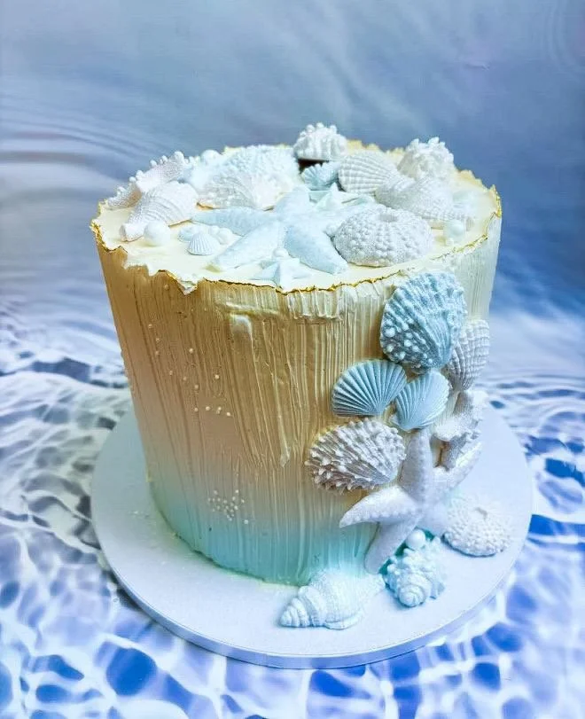 A tall ocean-themed decorated cake with seashell and coral decorations on top and around the sides, set against a blue and white watery background.