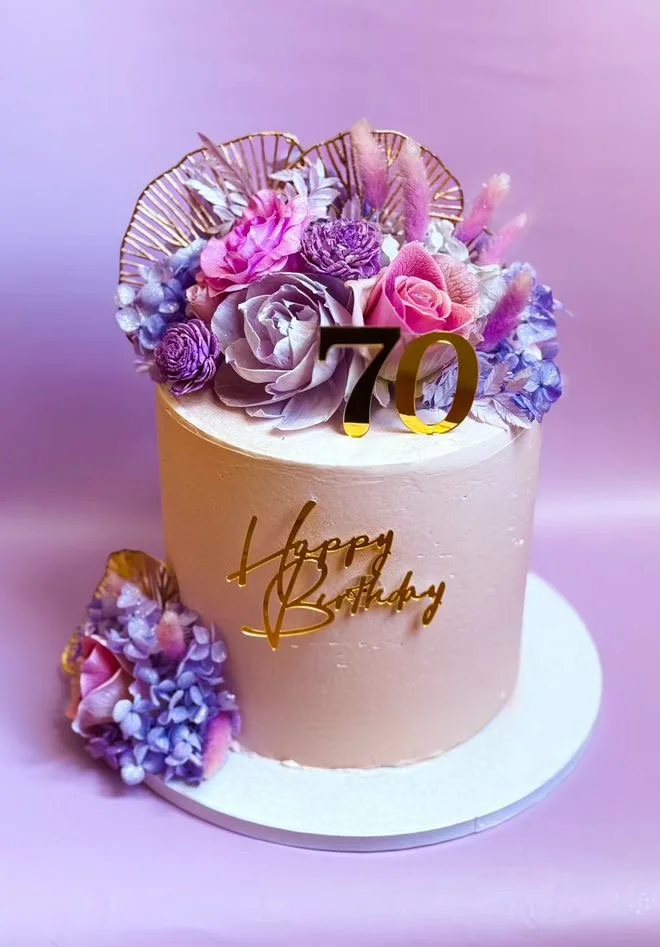A birthday cake decorated with pink and purple flowers, gold accents, and a "70" topper, with a gold "Happy Birthday" message on the front.