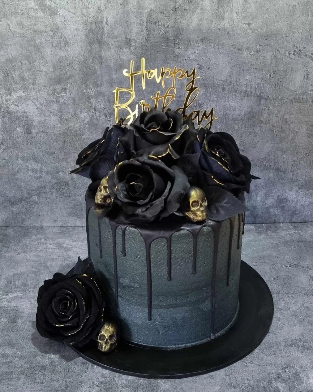 Black birthday cake with gold accents, decorated with black roses, small gold skulls, and a gold "Happy Birthday" topper, on a black cake board against a gray background.