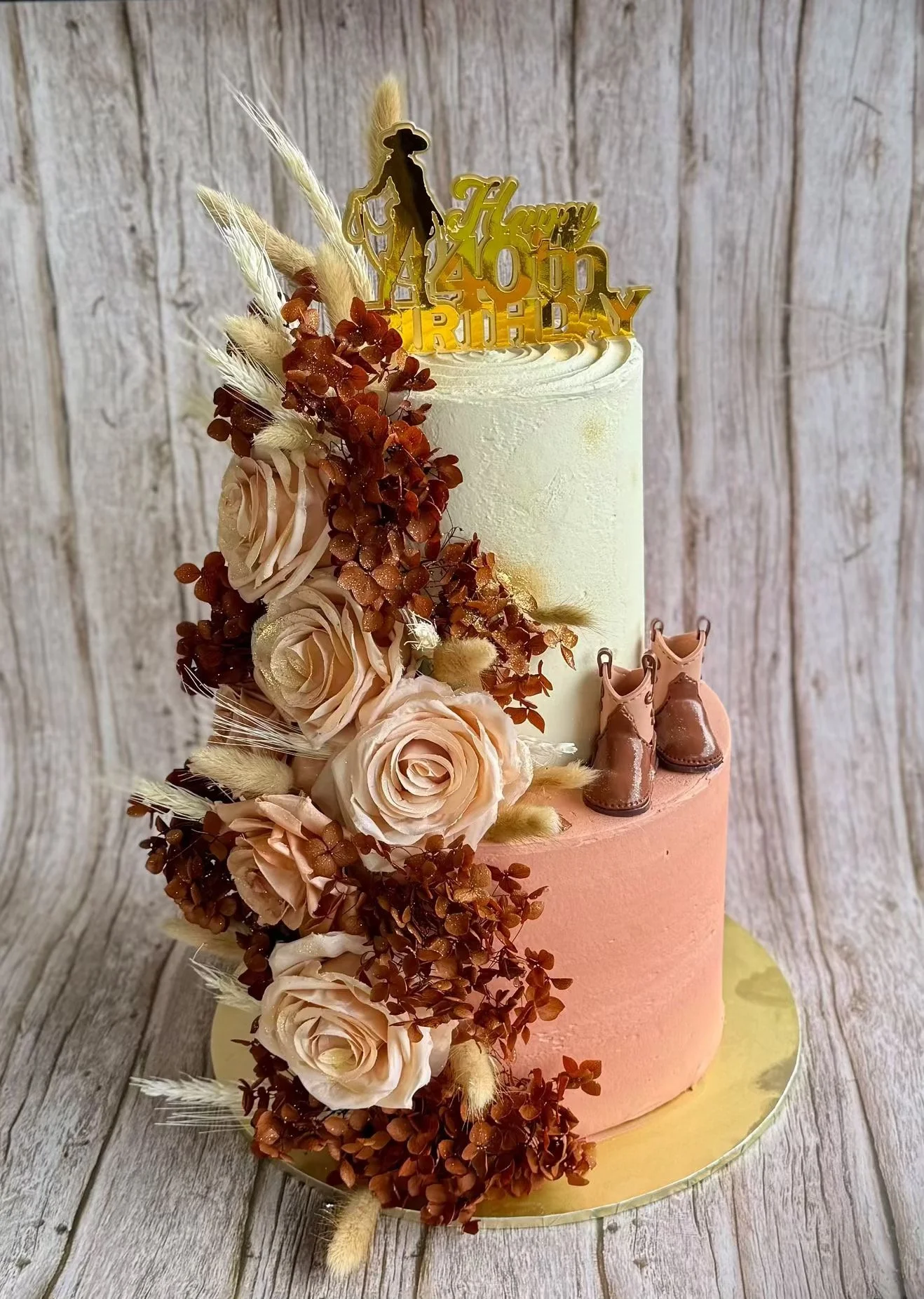 A two-tiered birthday cake with flower and cowboy boot decorations, featuring a gold 'Happy 40th Birthday' topper, set against a wooden background.