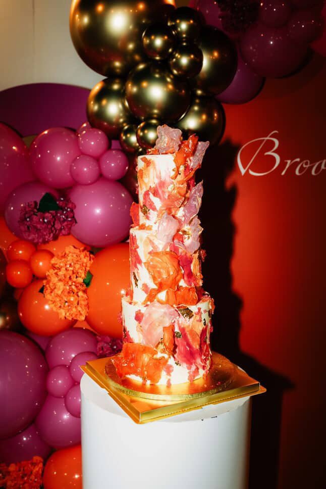 A tall, vertically stacked dessert with pink, orange, and white marbled decorations, sitting on a white pedestal with a gold square base, in front of a colorful balloon backdrop with shades of pink, orange, and gold.