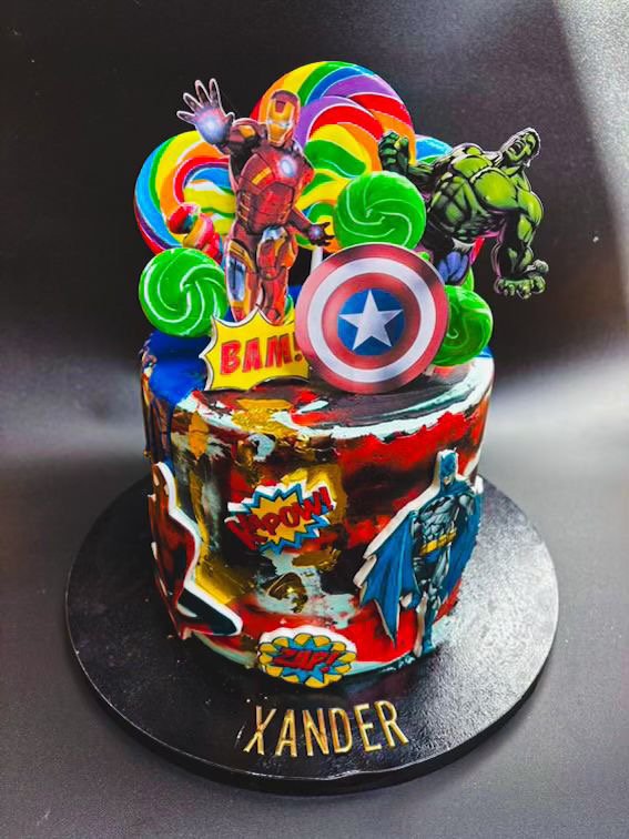 Decorated superhero-themed cake featuring Iron Man, Hulk, Captain America shield, and comic book style words like "BAM!" and "POW!"