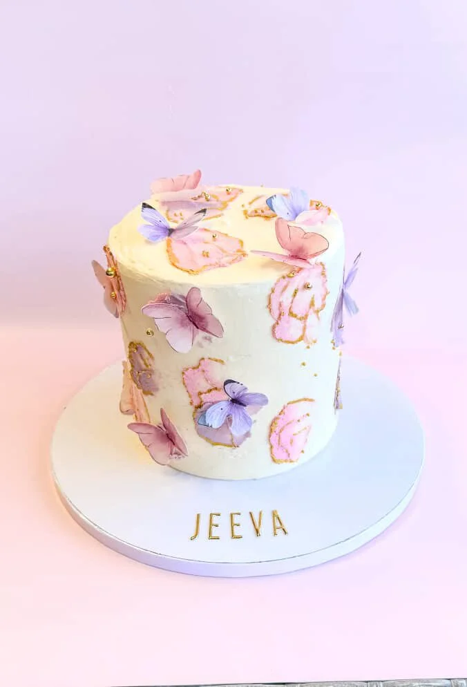 White cake decorated with pink and purple butterflies, gold accents, and pink frosting details, with the name 'JEEYA' in gold letters on the cake board.