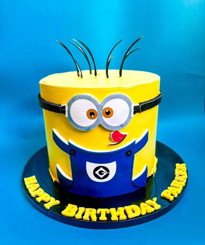 A Minions-themed birthday cake with yellow icing, adorned with Minions' goggles, eyes, and a smile, decorated with black hair made from wires, and a blue overall design, placed on a black cake board with yellow lettering saying 'Happy Birthday'. 