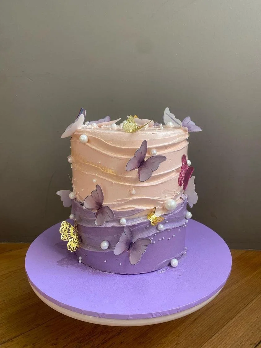 Two-tiered cake with pastel pink and purple buttercream, decorated with edible pearl sugar and purple, gold, and pink butterfly toppers, on a purple cake board.