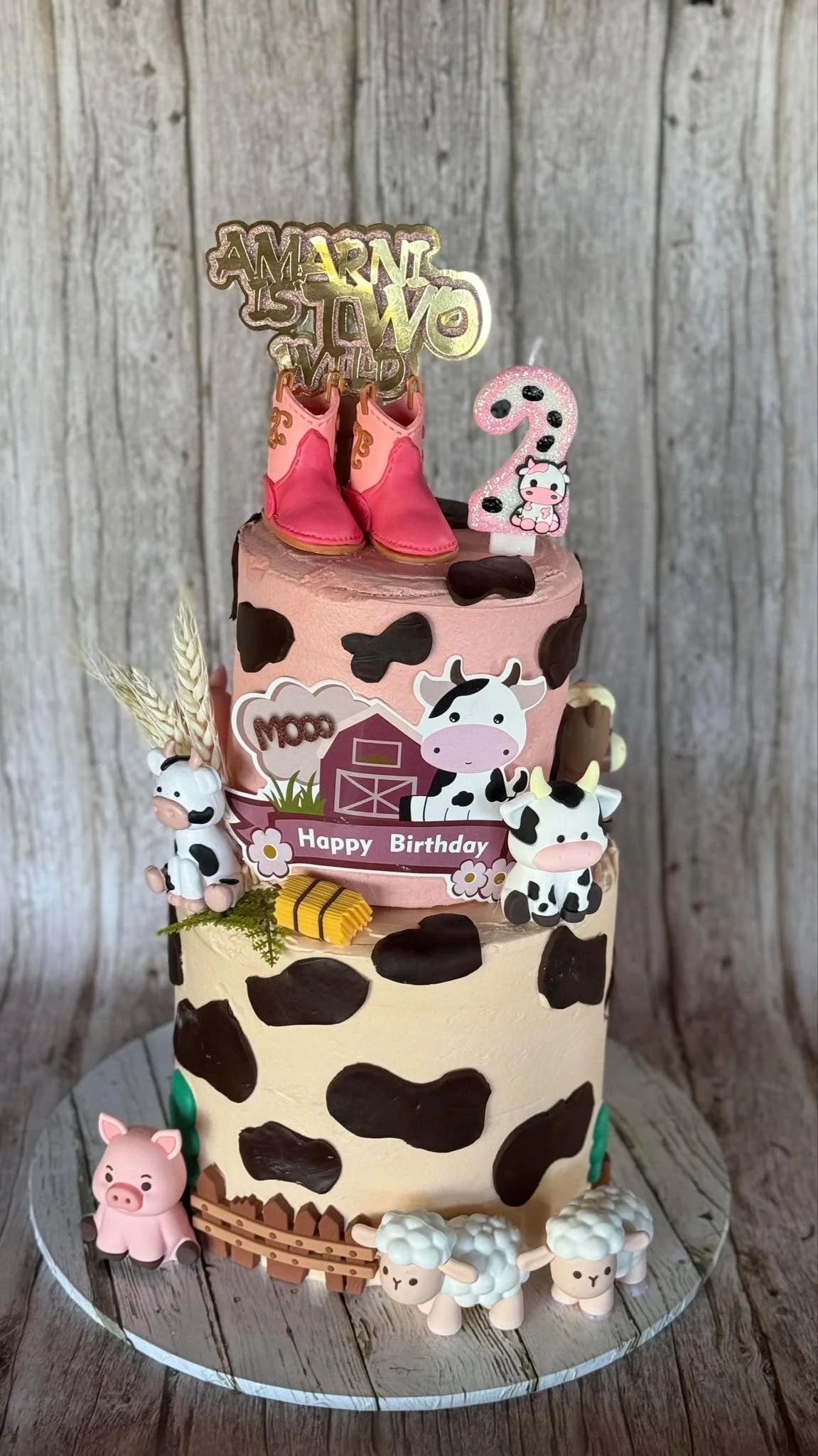 A multi-tiered cowboy-themed birthday cake decorated with cow print patterns, fondant farm animals, including cows, pigs, and sheep, and topped with pink cowboy boots, a birthday sign, and a number 2 candle with a cow design.