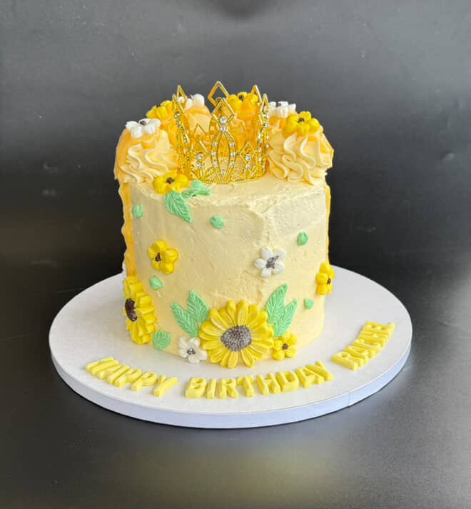 Yellow birthday cake decorated with sunflower and leaf-shaped icing, adorned with a gold crown-like topper made of edible decorations, placed on a white cake board with yellow letters spelling "HAPPY BIRTHDAY GABRIEL".