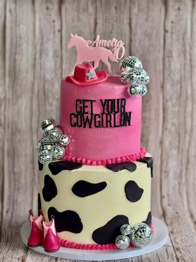 Two-tiered cake with cow print design on the bottom tier and pink frosting on the top tier. Decorated with small silver disco ball ornaments, a pink cowboy hat, pink cowboy boots, and a topper with a horse silhouette and the name Amelio. Written on t