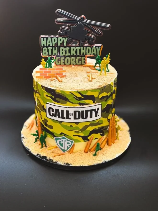 Two-tier birthday cake with Call of Duty theme, decorated with military figures, sand-like surface, and camouflage pattern. Topper includes a helicopter and a sign that reads "Happy 8th Birthday George".