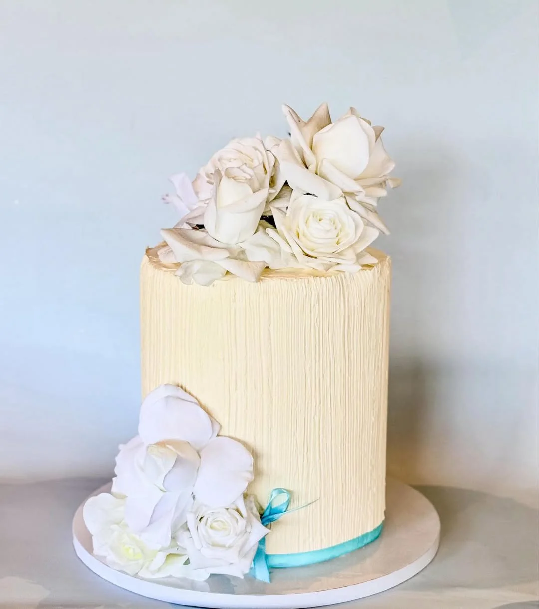 A tall, cylindrical cream-colored cake decorated with white roses and white petal-like decorations, with a blue ribbon around the base.