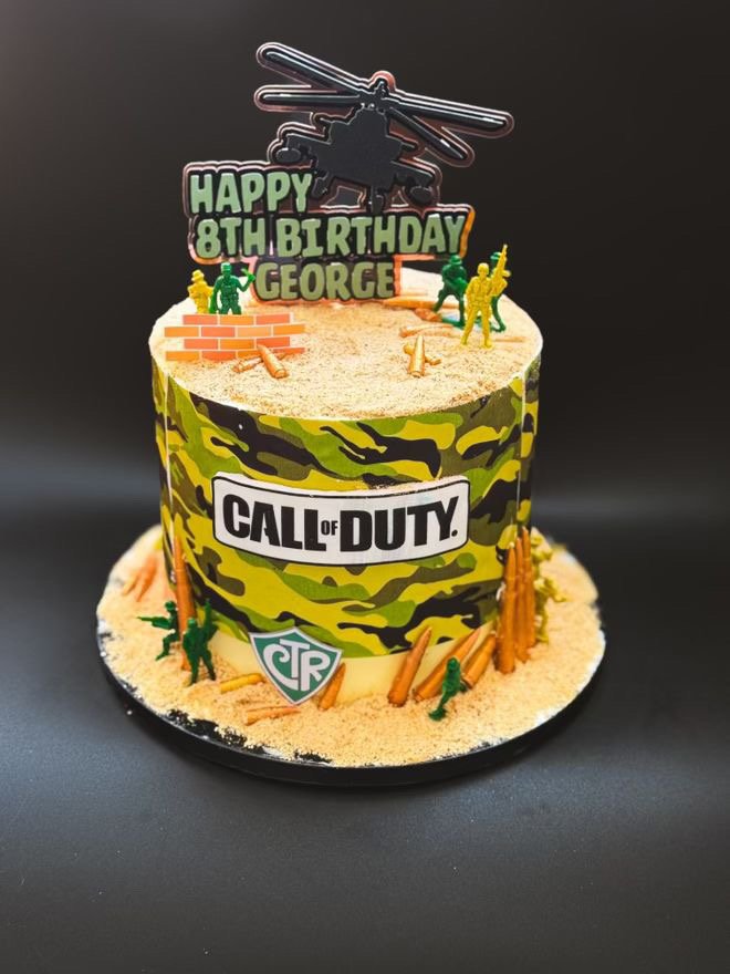 A two-tier birthday cake decorated with a Call of Duty theme, featuring a camouflage pattern, miniature toy soldiers, sand-like topping, and a battlefield scene with a helicopter and weapons, with a sign reading 'Happy 8th Birthday George' on top.