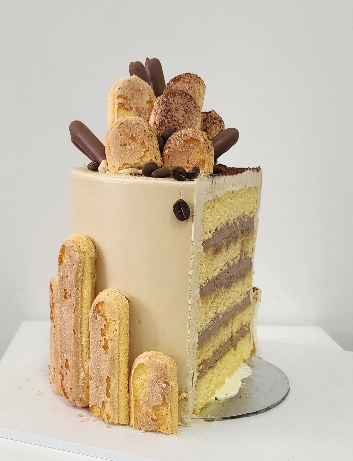 A layered vanilla cake with cream frosting, decorated with ladyfinger biscuits, chocolate-covered biscuits, coffee beans, and cocoa powder on top.