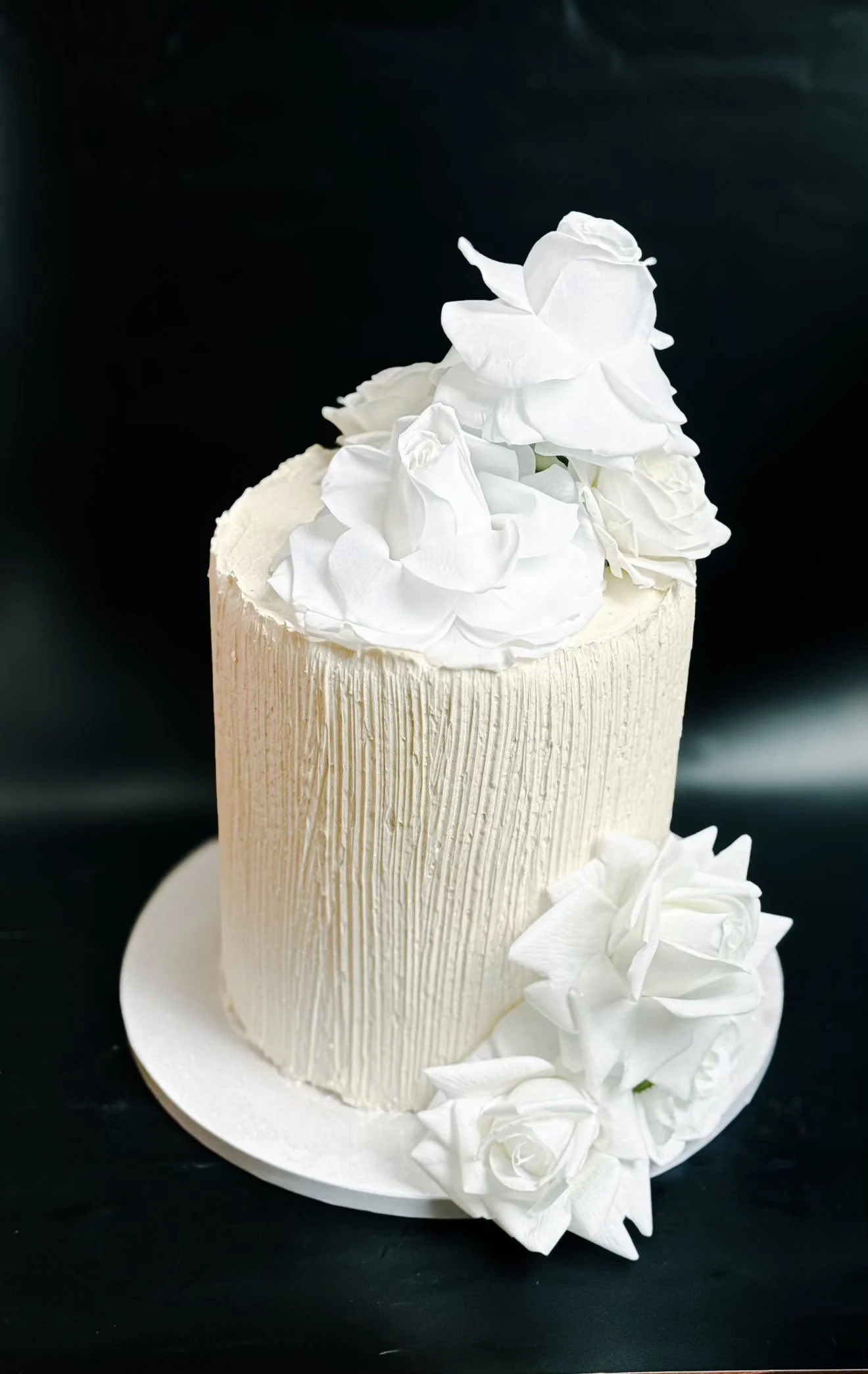 White wedding cake with textured vertical lines, decorated with white roses and petals, placed on a white cake board against a dark background.