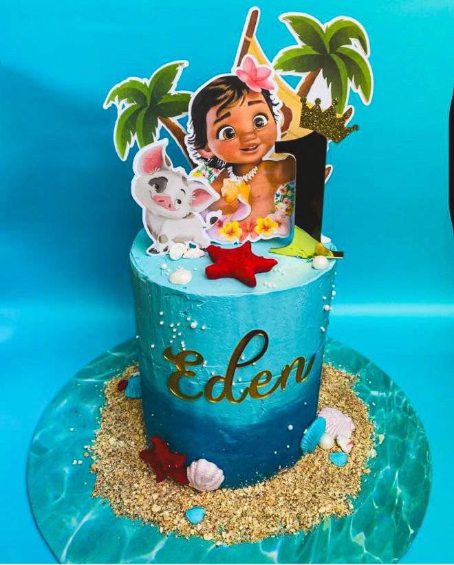 Blue ocean-themed birthday cake with edible decorations including a character from Moana, a pig with a flower, and tropical elements, featuring the name 'Eden' in gold lettering.