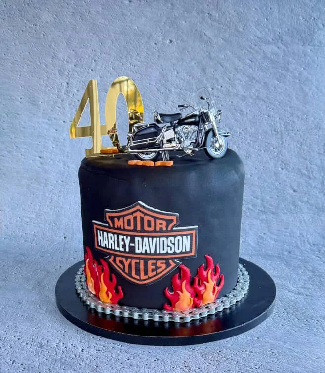 A Harley-Davidson themed cake decorated with a miniature motorcycle on top, a gold number 40, a Harley-Davidson logo, and red and yellow flame designs at the base.