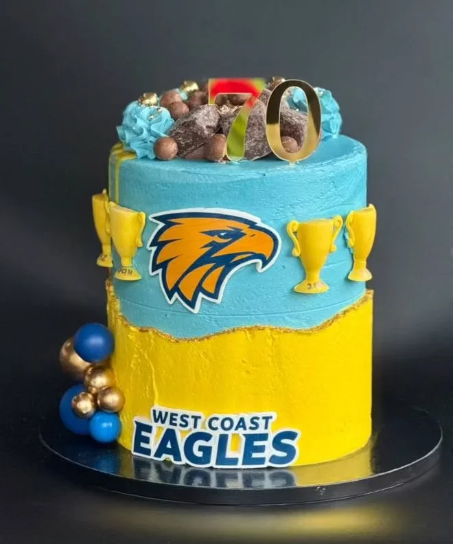 A two-tiered birthday cake with blue and yellow frosting, decorated with eagle logo, golden trophy toppers, and edible candies, celebrating 'West Coast Eagles' football team.