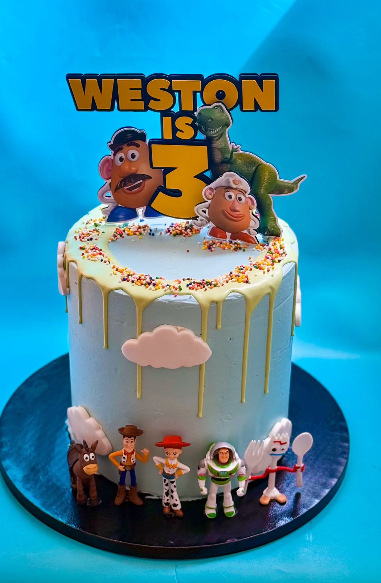 A Toy Story themed birthday cake with figures of Woody, Buzz Lightyear, and other characters at the base, and a large sign reading 'Weston is 3' with cartoon character toppers and colorful sprinkles.