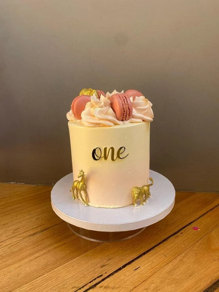 A white birthday cake with the word 'one' in black and gold text. The cake is decorated with pink macarons, swirls of pink frosting, and gold elephant figurines on the sides. It sits on a white cake base on a wooden surface.