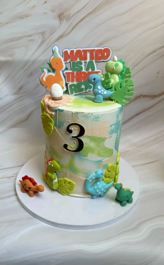 A birthday cake decorated with a dinosaur theme, featuring small colorful dinosaur figurines, leafy decorations, and a topper with the phrase 'MATTED IS A THE RE'. The cake has a large black number 3 on it, indicating a third birthday.