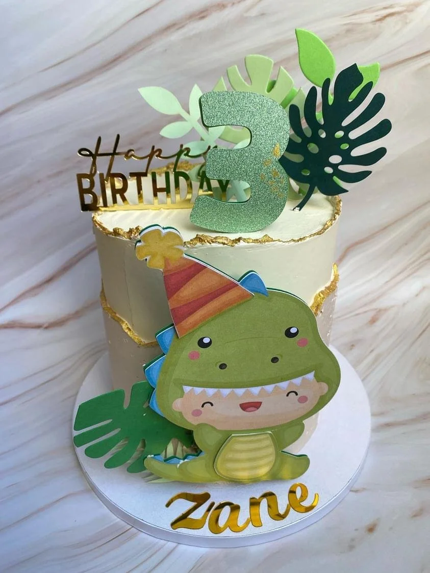 A birthday cake decorated with a green dinosaur figure, a large glittery number '3', tropical leaves, and a 'Happy Birthday' topper. The dinosaur wears a striped party hat and the cake is on a white marble surface.