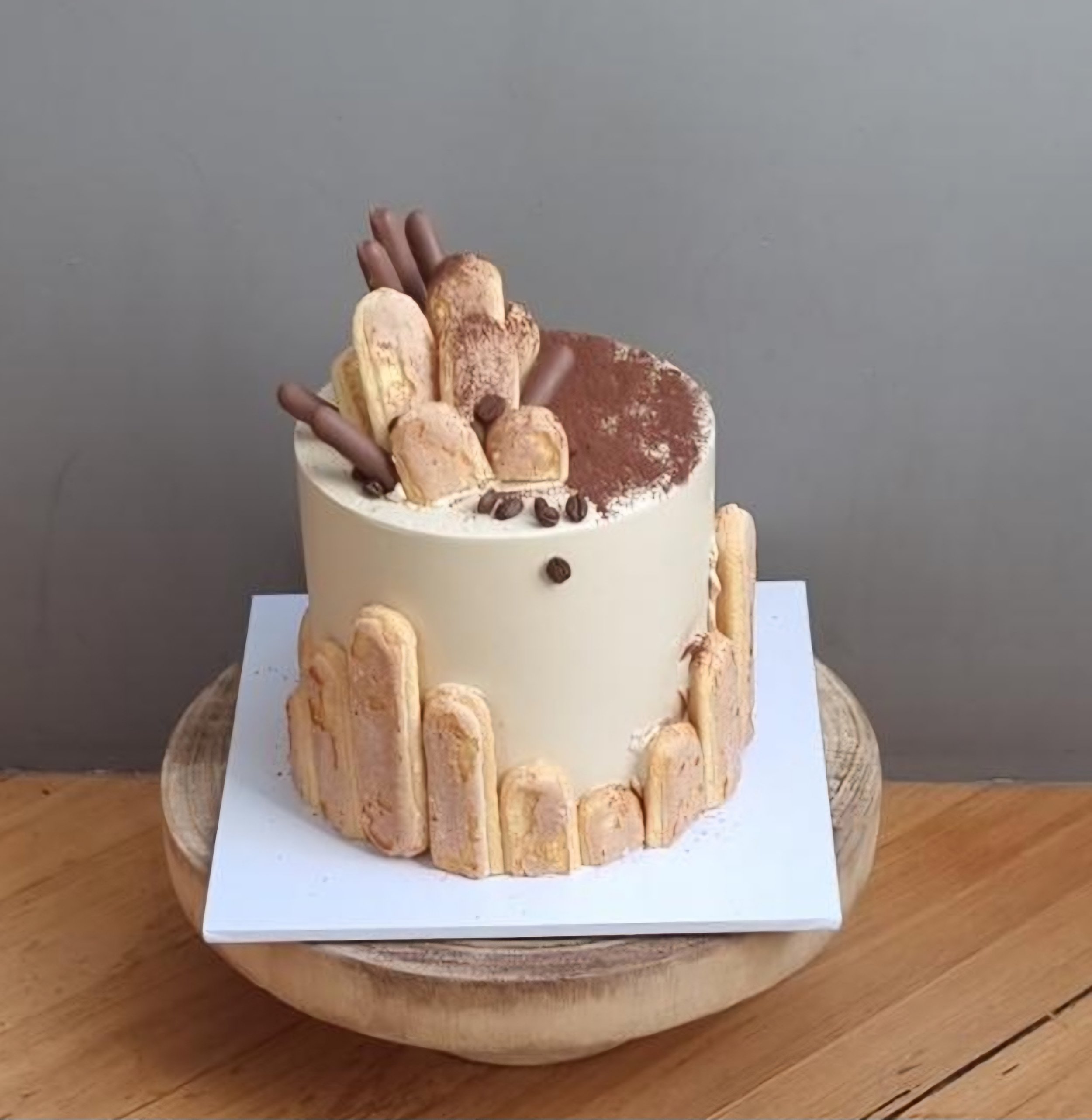 A cake topped with ladyfingers, chocolate decorations, and cocoa powder, on a wooden table.
