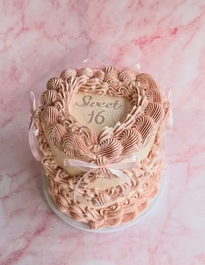 Pink and white heart-shaped birthday cake with piped frosting and a ribbon, inscribed with 'Sweet 16' on top.