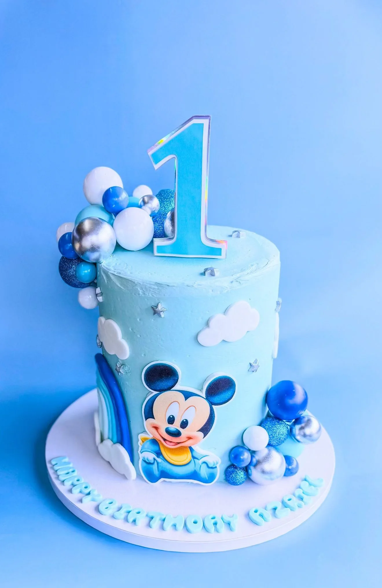 Blue birthday cake with Mickey Mouse decoration, blue and silver balloons, a large number one topper, and fondant clouds on a white base with birthday messages.