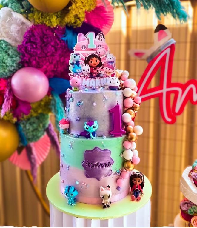 Colorful three-tiered birthday cake decorated with small toys, flowers, and candies, celebrating a child's first birthday, with a backdrop of balloons and decorations.