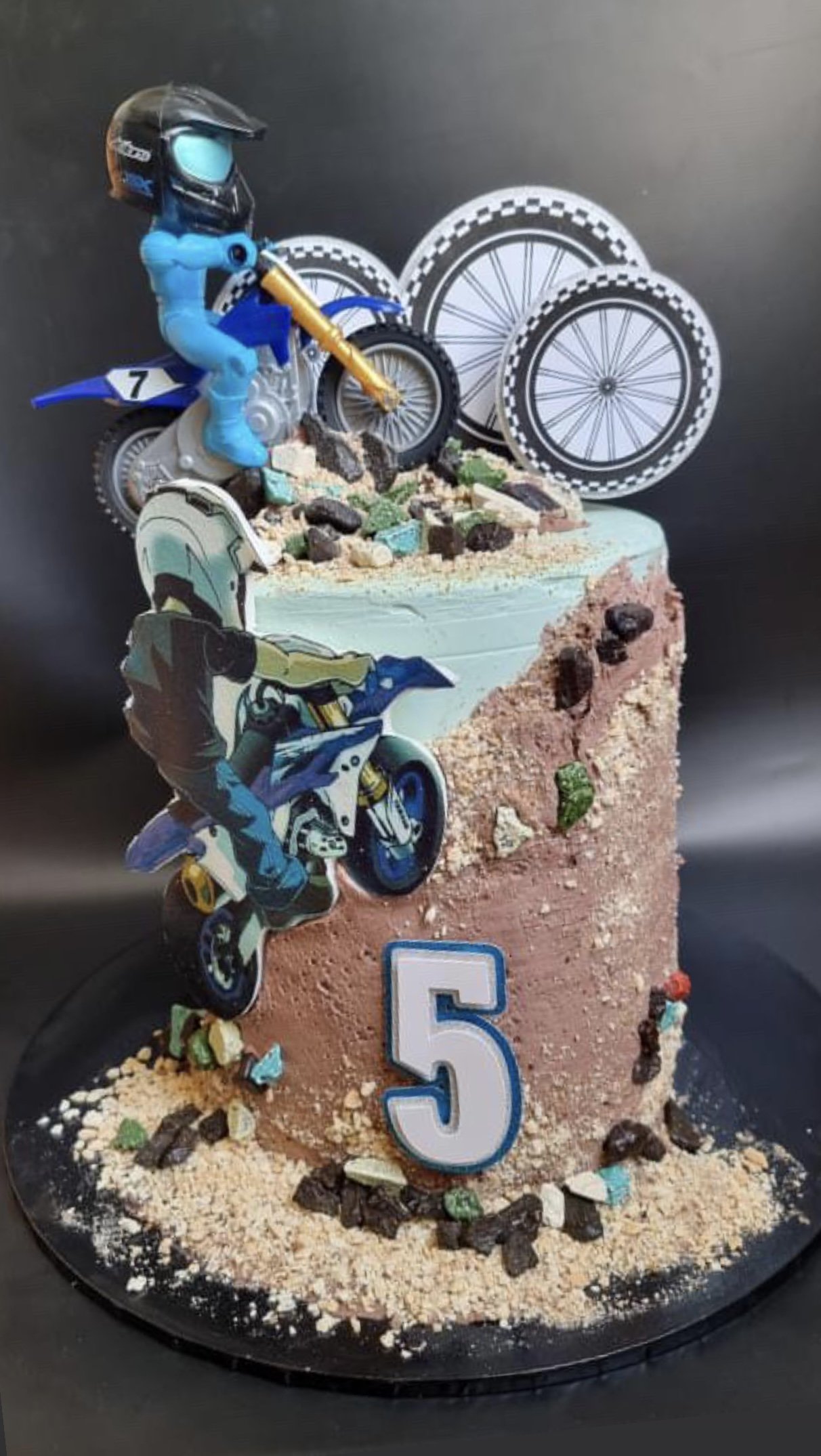 Cake decorated with motocross theme, featuring cutouts of motocross riders and wheels, sand-like cake decorations, and a number five