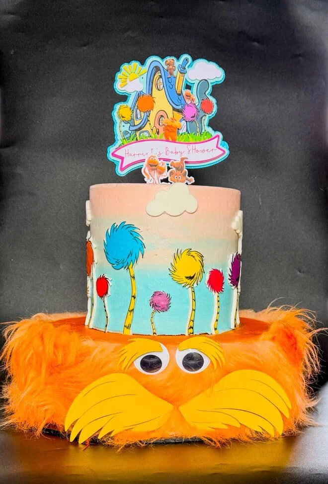 A cake decorated with colorful jungle animals and trees, topped with a "Happy 1st Baby Shower" sign, placed on a furry orange base with googly eyes to resemble a lion's face.