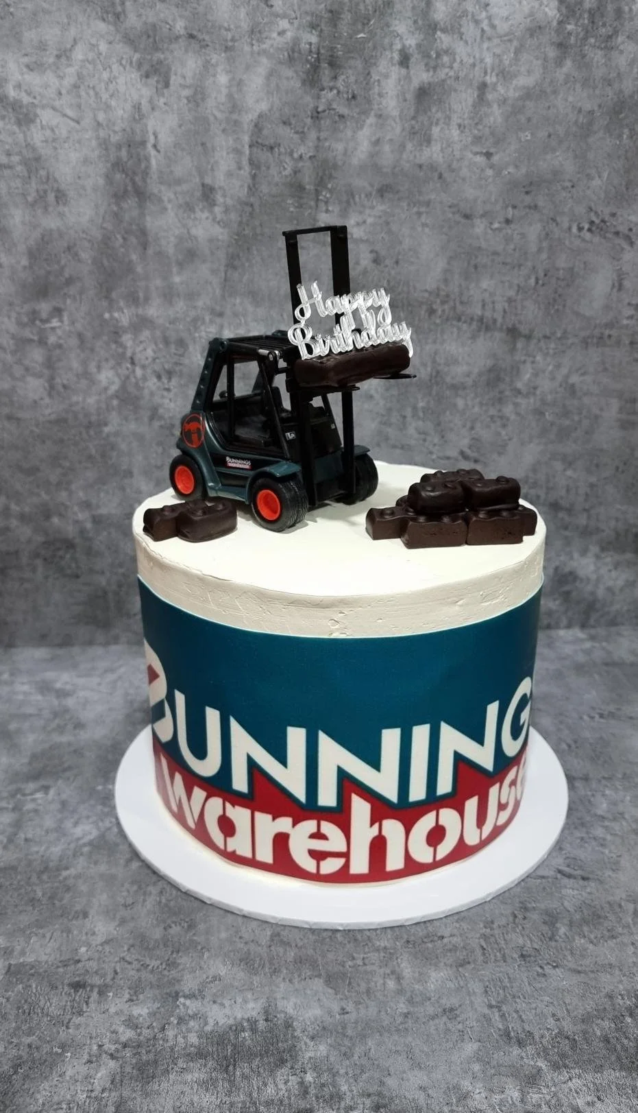 Celebration cake with a 'Happy Birthday' topper and a toy forklift on top, decorated with chocolate treats, featuring a blue and red 'Running Warehouse' logo on the side.