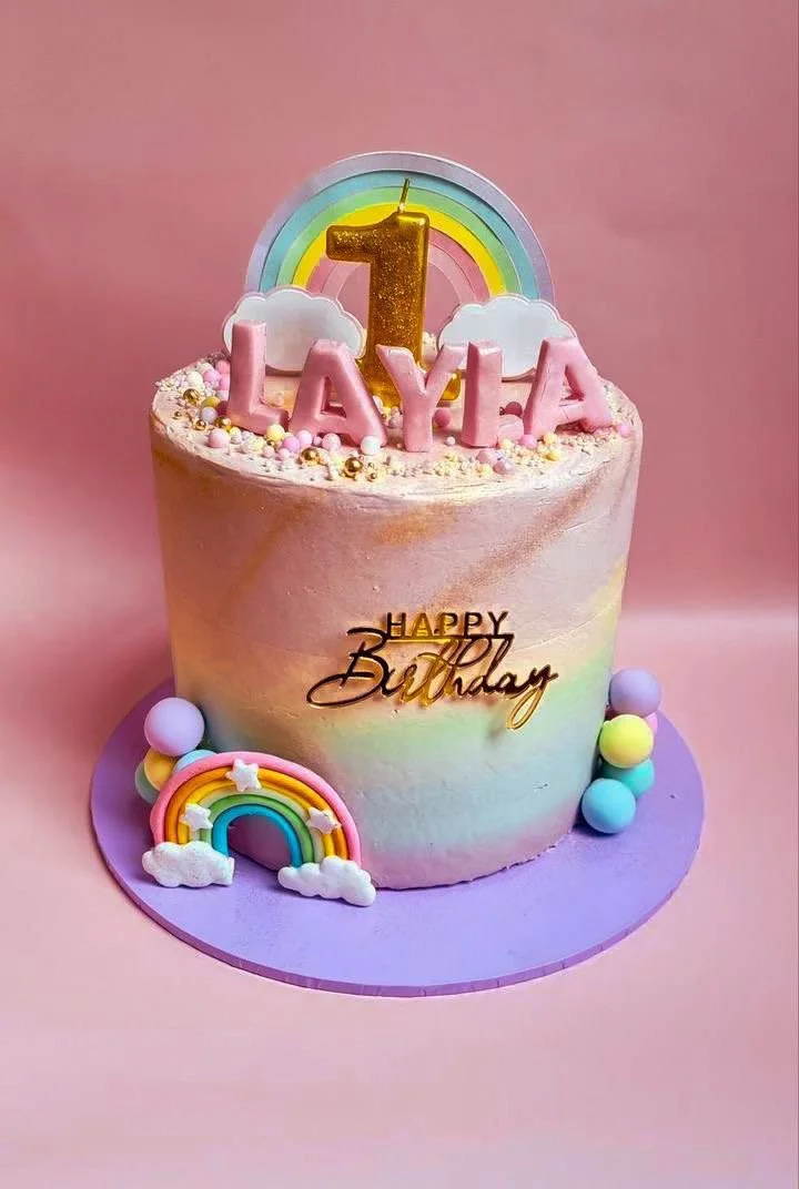 A colorful birthday cake with pink frosting, rainbow decorations, and pastel-colored balls. It has a gold candle shaped like the number one, and pink letters spelling "LAYA" on top. The cake has a "Happy Birthday" topper and is set on a purple cake b