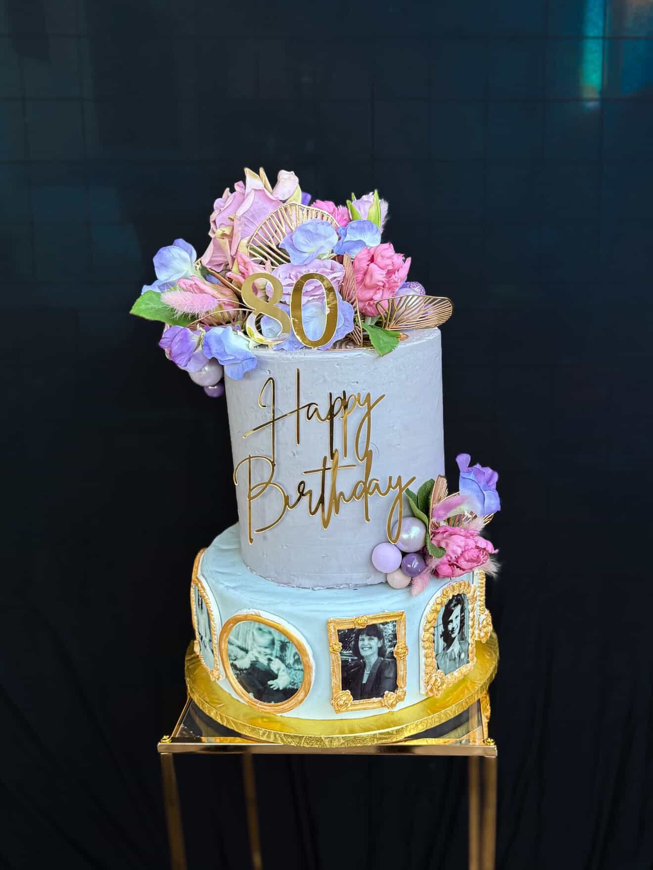 A two-tiered birthday cake decorated with flowers, gold accents, and black-and-white photos. The top tier has a gold "80" topper and the words "Happy Birthday" written in gold cursive. The cake is on a gold stand with a black background.