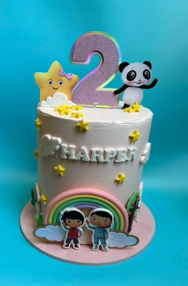 A birthday cake decorated with a large purple number 2, a star with a bow, a panda plush toy, and rainbow and cloud decorations, celebrating a child's second birthday.