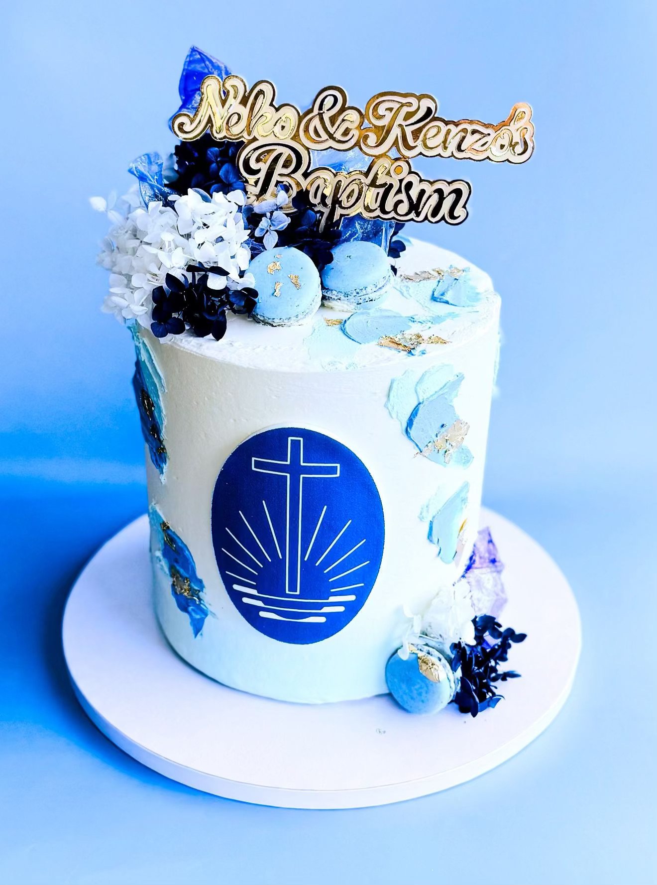 A white birthday cake decorated with blue and white flowers, macarons, and edible gold leaf, featuring a religious symbol and a gold-colored cake topper that reads 'Neko & Kenzos Baptism' in gold and black lettering.